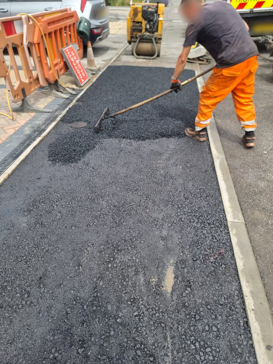Worker raking tarmac between a block-paved drive and the kerb line, with barriers and a roller behind