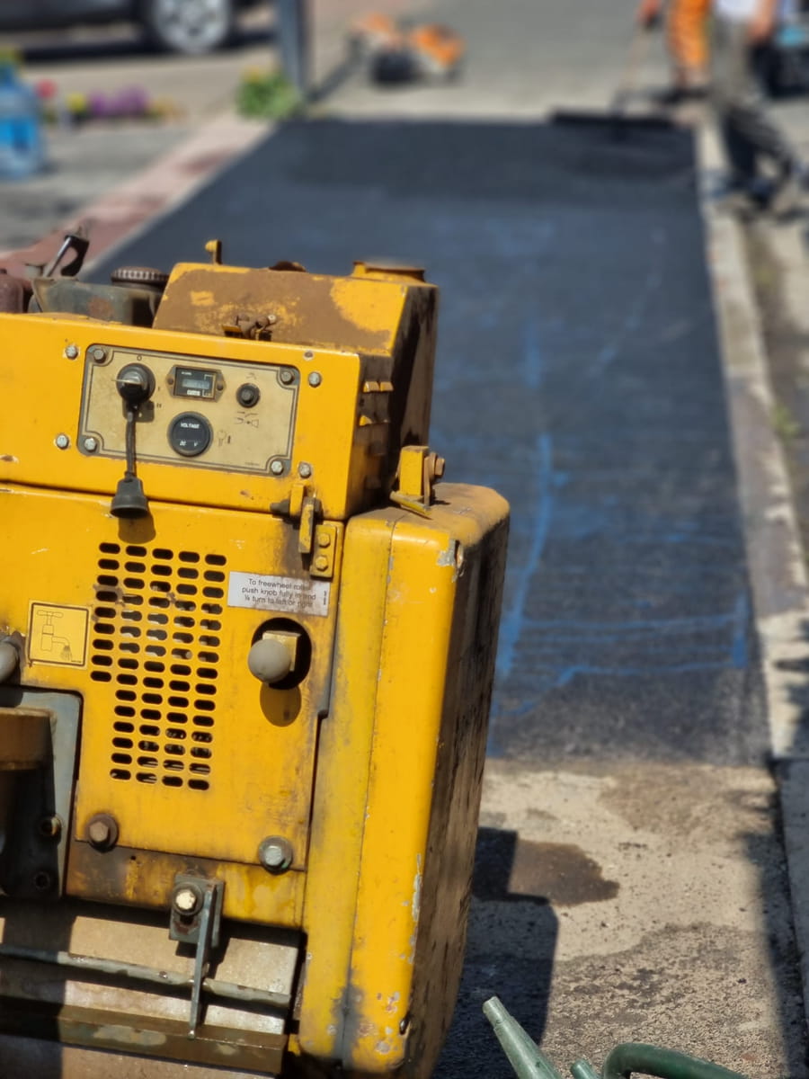 Yellow roller on freshly laid tarmac beside a kerb