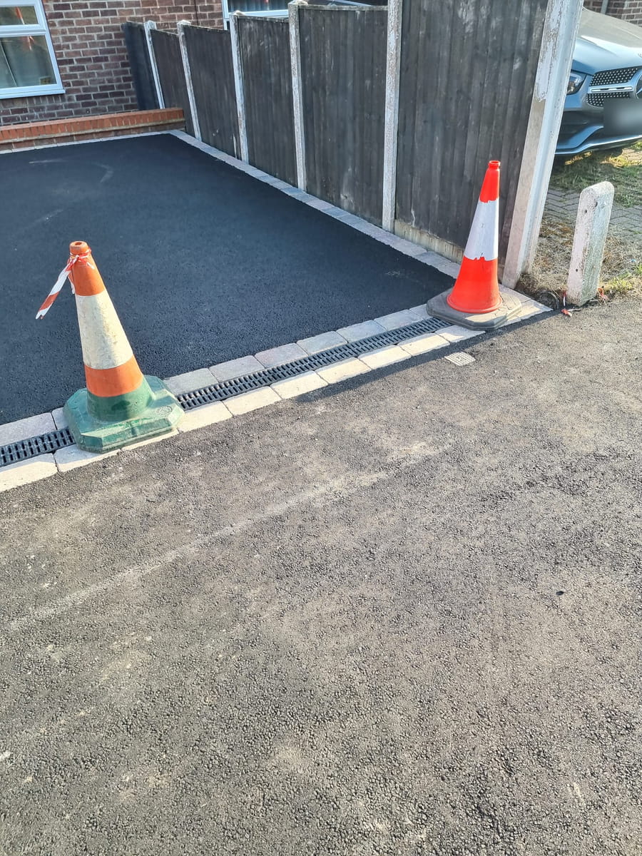 New tarmac driveway with a channel drain and block edging where it meets the pavement, traffic cones still out